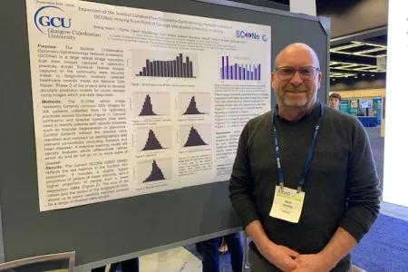 An image of Niall Strang, a white male wearing a dark jumper with an ARVO lanyard round his neck, standing next to his poster at a conference.