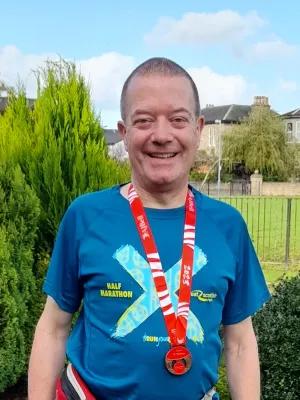 A man wearing a blue t shirt and a Glasgow half marathon medal around his neck photographed standing outside on a sunny day.